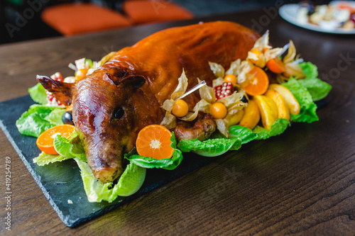 Restaurant menu. Baked suckling pig whole on a stone decorative board with fruit and physalis.  View from the head.