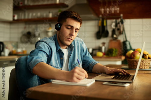 Young man writing notes while working on a computer at home.