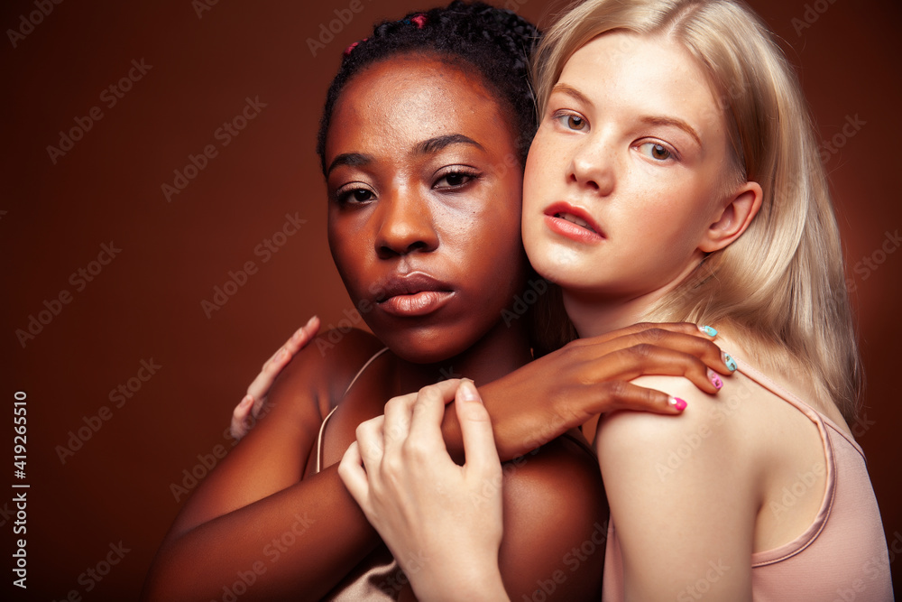 two pretty girls african and caucasian blond posing cheerful together on brown background, ethnicity diverse lifestyle people concept