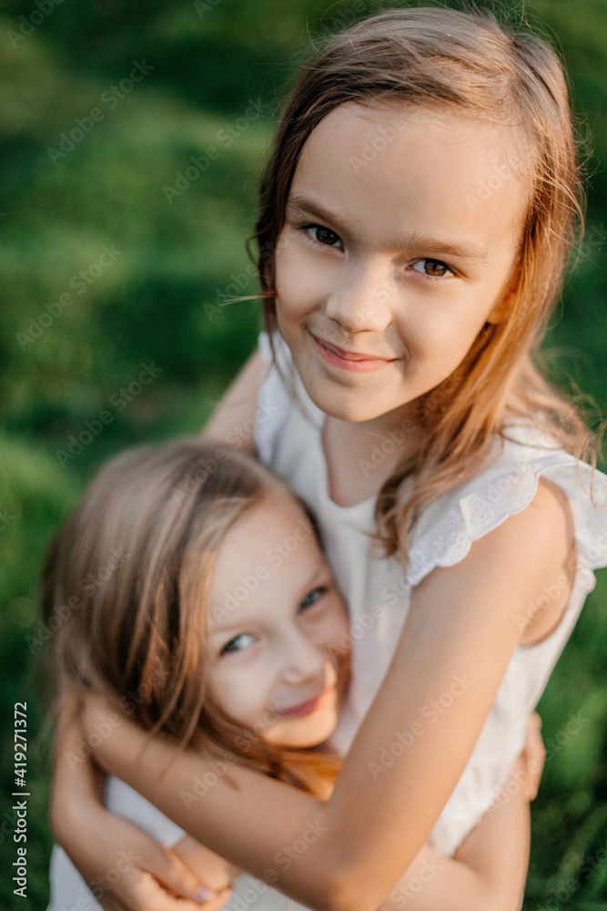 Happy children in white dresses smile standing on the grass in summer. Portrait of two sisters. Children's friendship Happy children in white dresses smile standing on the grass in summer. Portrait of two sisters. Children's friendship