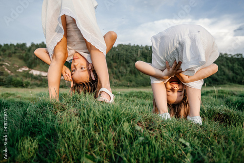 Upside down photo of happy children in white dresses play on the grass and smile in summer. Funny portrait of two sisters. Children's friendship