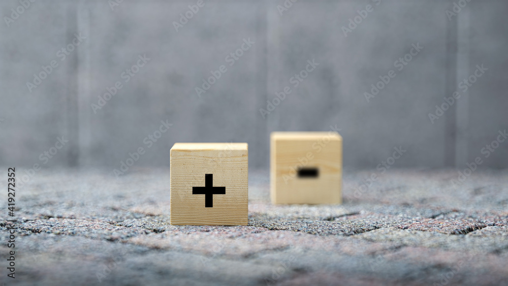 Plus and Minus icons on wooden cubes with selective focus to the Plus over a grey background with copyspace in a conceptual image Plus and Minus icons on wooden cubes with selective focus to the Plus over a grey background with copyspace in a conceptual image