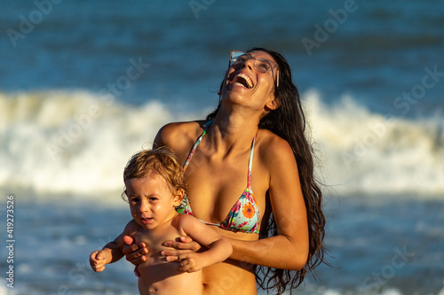 MÃE SORRINDO E SEGURANDO FILHO NO COLO EM UMA PRAIA BRASILEIRA.