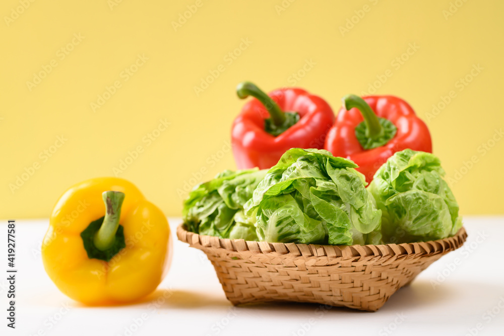 Fresh cos lettuce and bell peppers in a basket on white and yellow background Fresh cos lettuce and bell peppers in a basket on white and yellow background