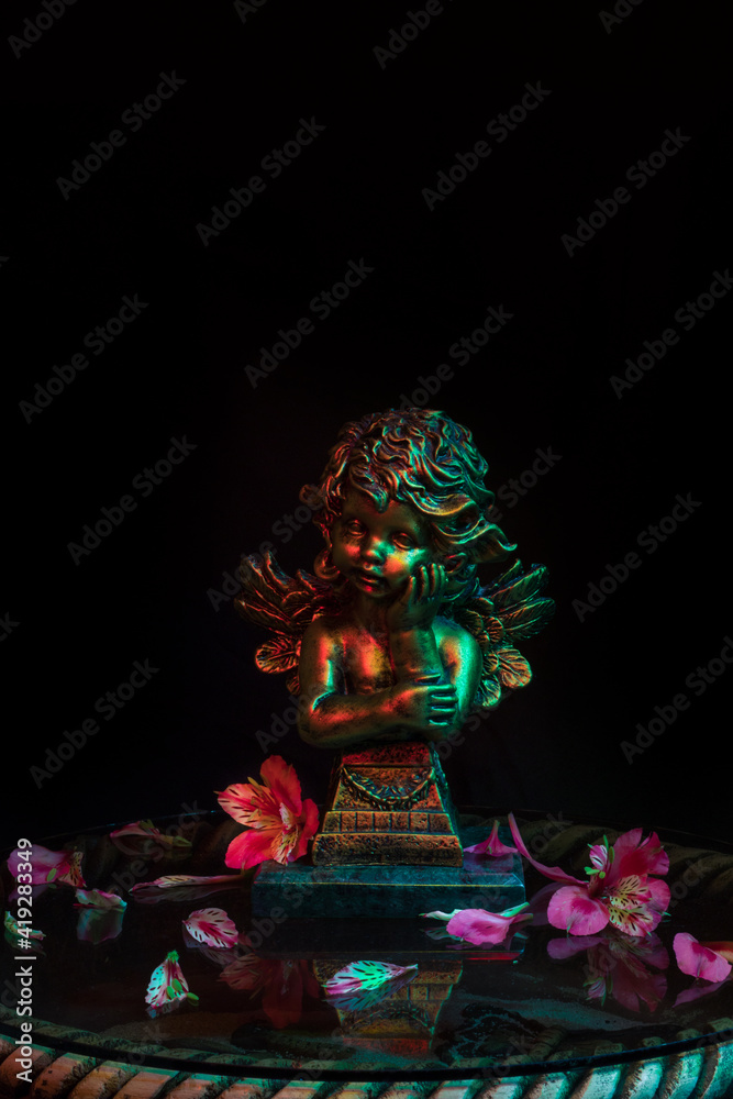 Golden angel sculpture adorned with flowers on a glass with warm and cold lights on a dark background