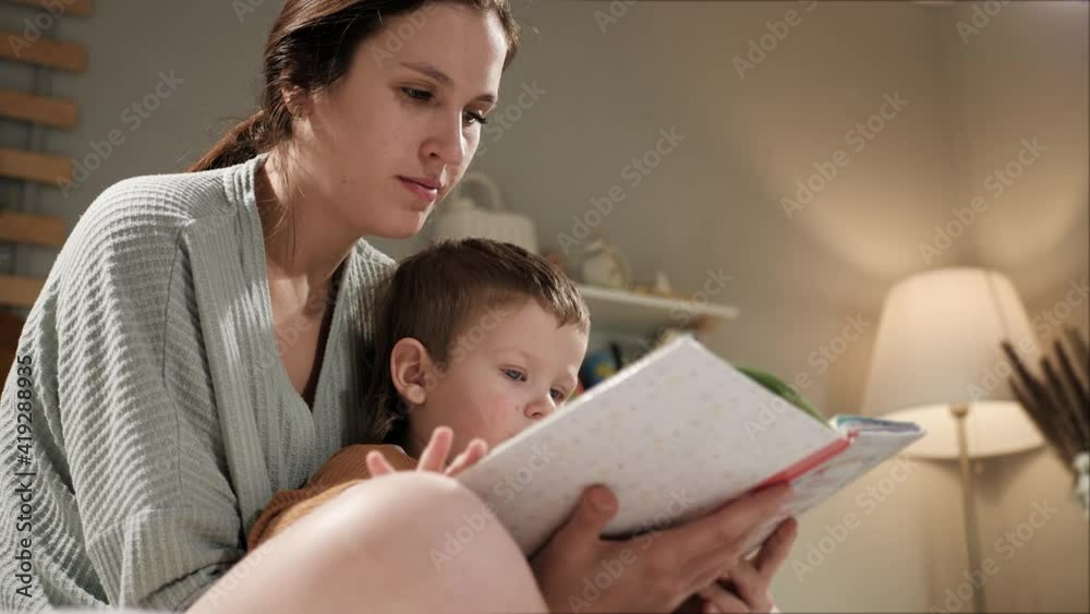 Baby and book, mother and child reading. Woman mom and 2-3 year old boy sit on bed in pajamas in bedroom in evening or night and hold book in hands, looking and reading, turn page. Slow motion Baby and book, mother and child reading. Woman mom and 2-3 year old boy sit on bed in pajamas in bedroom in evening or night and hold book in hands, looking and reading, turn page. Slow motion