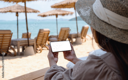 Mockup image of a woman holding mobile phone with blank desktop screen while sitting on the beach
