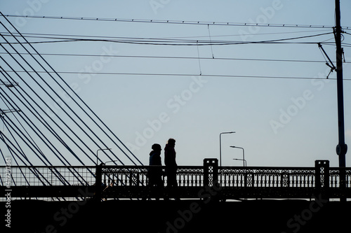 The silhouettes of two people crossing a suspension bridge on foot. Photo during the day. © samy