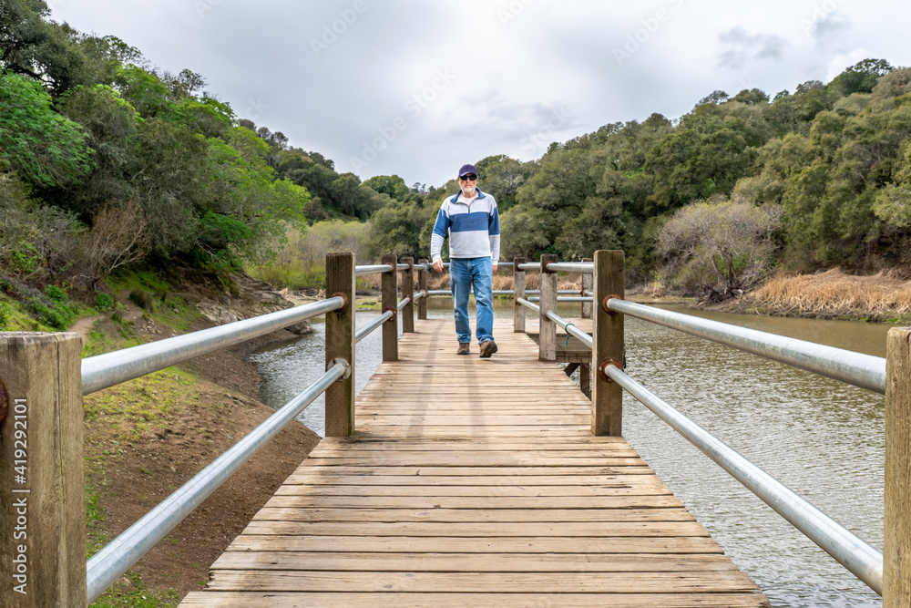 A man, visitor, hiker walking toward small on a pier or a wharf over a small lake with tree covered trees on both sides and cloudy sky overhead, Waterdog Lake, Belmont, California