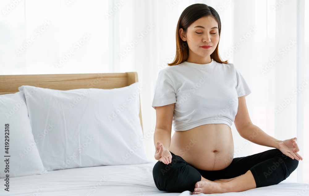 young pretty Asian pregnant woman sitting on a bed doing exercise delightfully by Yoga. She is smiling and looking at a camera. Healthy happy mother concept young pretty Asian pregnant woman sitting on a bed doing exercise delightfully by Yoga. She is smiling and looking at a camera. Healthy happy mother concept