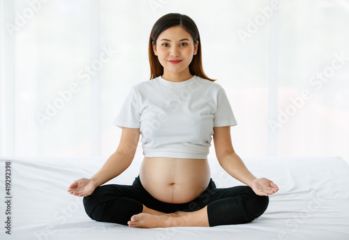 young pretty Asian pregnant woman sitting on a bed doing exercise delightfully by Yoga. She is smiling and looking at a camera. Healthy happy mother concept