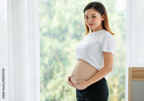 portrait of a good-looking Asian pregnant woman dress casually standing and touching her belly with love and happiness while looking and smiling at a camera Free Download portrait of a good-looking Asian pregnant woman dress casually standing and touching her belly with love and happiness while looking and smiling at a camera - GFXLoad