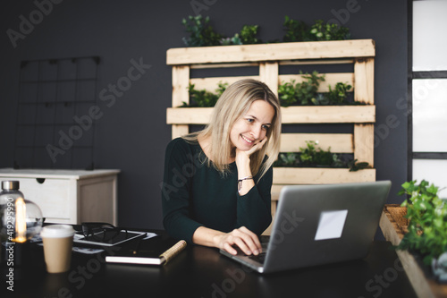 pretty blond young woman is sitting in an ecological office with lots of plants and is working on her laptop and is wearing a green sweater, concept sustainability and environment today