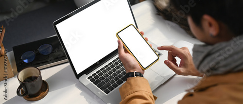 Horizontal photo of young man sitting in front of laptop computer and holding smart phone with blank screen.