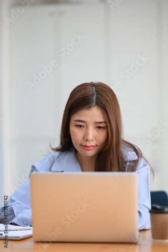 Portrait of young female office worker working on laptop computer while sitting at office desk.