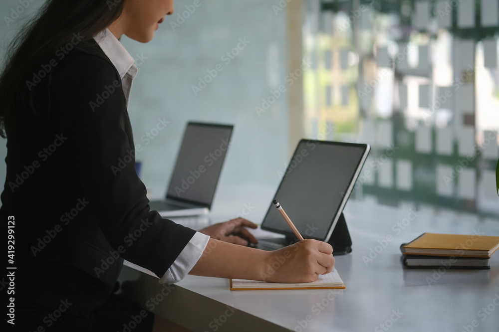 Cropped shot of businesswoman working with computer tablet and writing information on notebook at office desk.