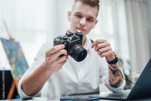 Professional photographer with tattooed arm looks at photocamera he holding it. Hipster guy wearing formal clothing working in office.