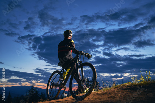 Back view of young man in cycling suit sitting on bicycle under beautiful night sky. Male bicyclist in safety helmet resting on hillside road under blue cloudy sky while riding bicycle in the evening.