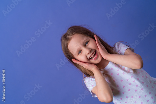 little girl 5 years old with blonde hair in a T-shirt with colored polka dots stands smiling and holding her hands to her face on purple background. model is having fun and feels overwhelmed, amazed.