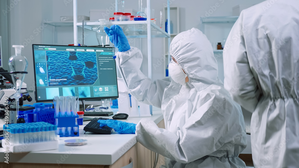Scientist nurse in ppe suit taking notes on computer looking at test tube in laboratory. Chemists examining vaccine evolution using high tech for research in treatment development against covid19 Scientist nurse in ppe suit taking notes on computer looking at test tube in laboratory. Chemists examining vaccine evolution using high tech for research in treatment development against covid19