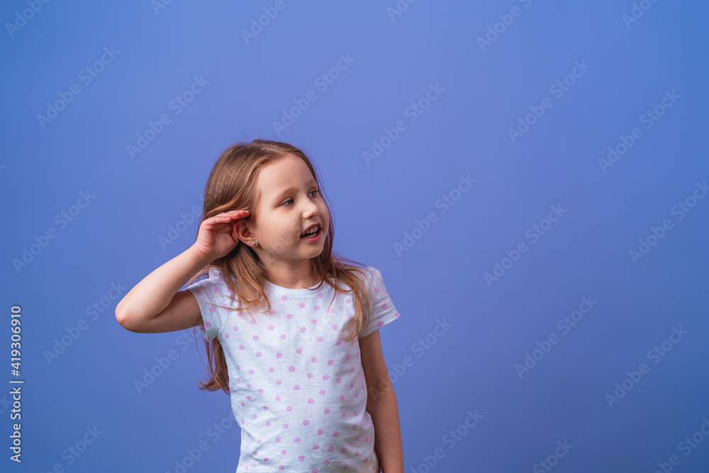 charming little girl smiles with her hand over her ear while listening and hearing rumors or gossip on a purple isolated background. The child is eavesdropping or hard of hearing