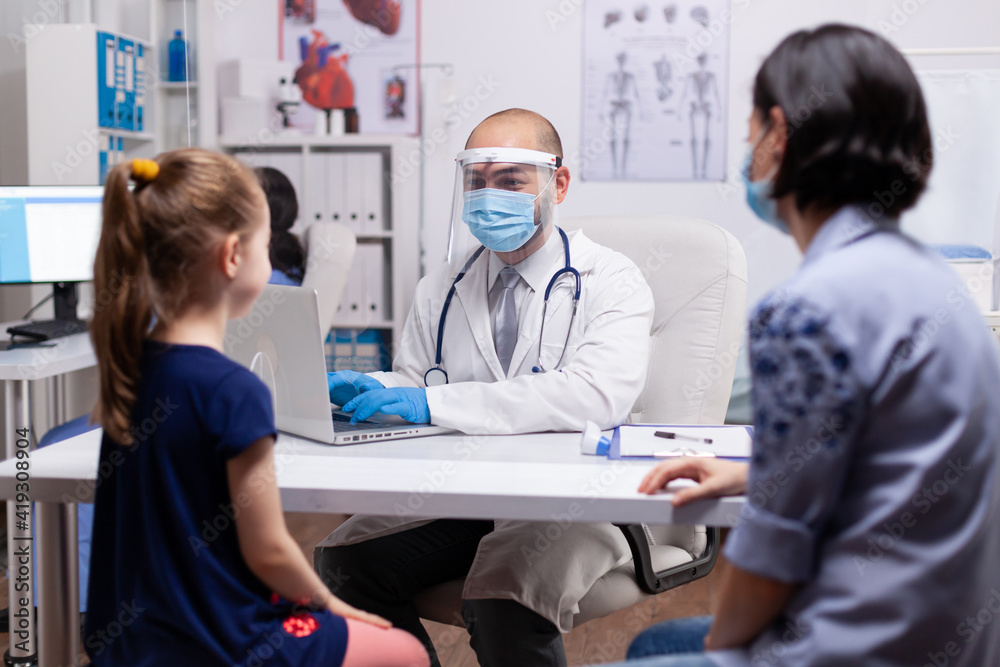 Pediatrician wearing protection mask and visor against covid-19 virus consulting child treating diagnostic of disease in new normal medical clinic. Therapeutist talking with mother during examination Pediatrician wearing protection mask and visor against covid-19 virus consulting child treating diagnostic of disease in new normal medical clinic. Therapeutist talking with mother during examination
