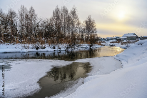 Fototapeta Naklejka Na Ścianę i Meble -  A winter evening falls over the village, painting the sky yellow. The sun is reflected in the unfrozen water, sparkling and shining brightly on the surface of the river. Far away wooden houses 