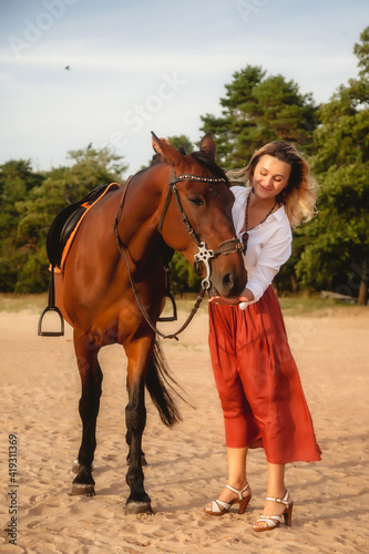 Cute happy young woman with horse in summer beach by sea. Rider female drives her horseback in nature on evening sunset light background. Concept of outdoor riding, sports and recreation. Copy space