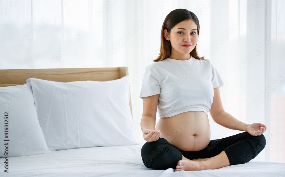 young pretty Asian pregnant woman sitting on a bed doing exercise delightfully by Yoga. She is smiling and looking at a camera. Healthy happy mother concept