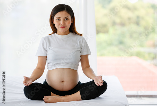 portrait of a beautiful Asian pregnant woman dressed casually sitting on a white bed close her eyes doing meditation after a workout by yoga in a bedroom at home