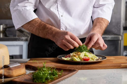 Professional Chef-cook Decorating Dish In Restaurant Kitchen Alone. Man In White Apron Makes Finishing Touch On DIsh, Adding Some Greens. Culinary, Restaurant, Gourmet Concept