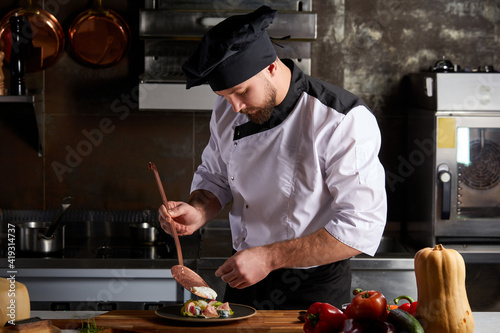 Chef adding piquancy to dish, finishing with flavouring of meal on plate, wearing cap and apron, cook alone in restaurant kitchen