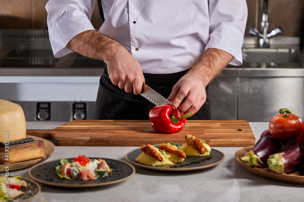 Slicing sweet red pepper on wooden cutting board, professional cook in white uniform preparing salad. Female hand cuts capsicum with knife. restaurant staff at work