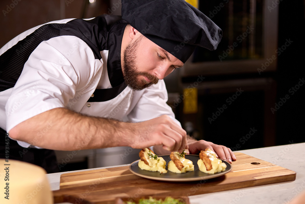 Chef in cap finishing dish on plate with dish dressing and almost ready to serve at table, professionally cooked and decorated meal for restaurant visitors
