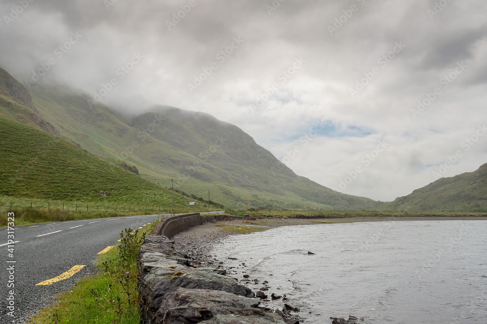 Small road in Connemara between a lake and mountains, Low cloudy sky. Ireland.
