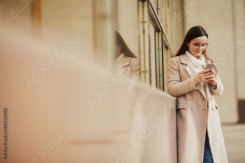 Smiling girl wearing beige coat and eyeglasses standing near building in the city street and uses her phone. 
