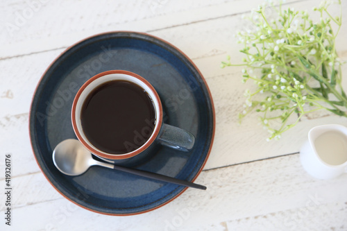 Coffee in a blue cup on a white wooden table. With a modern coffee spoon and milk pitcher, and butterweed flowers.