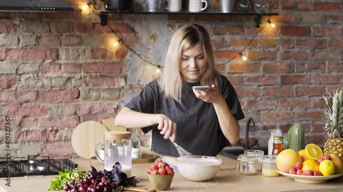 skilled culinary blogger holds smartphone and makes video of mixing dish products in bowl with metal whisk on brown wooden table in loft style interior against aged brick wall and garland Medium shot photo