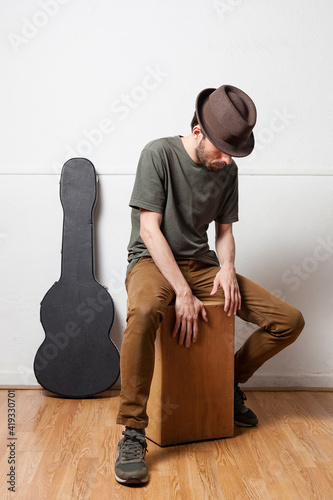 White spanish man wearing hat playing peruvian cajon on a white background with guitar case and wooden floor. Flamenco passion concept. photo