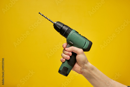 Green cordless battery powered drill on yellow background, cropped male hands holding tool for repair and building construction. copy space for advertisement