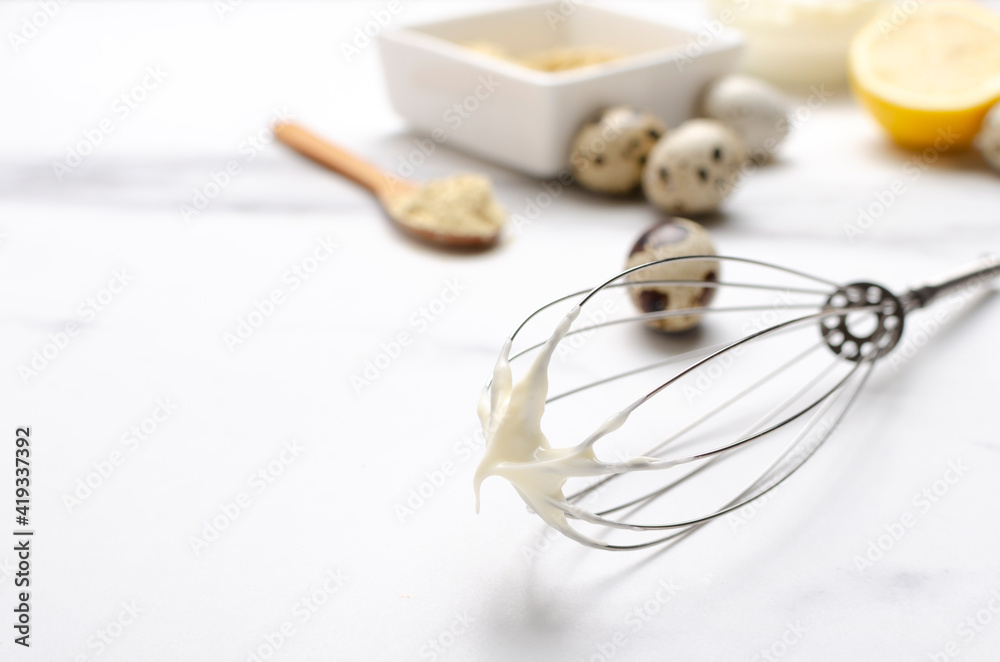 Metal whisk and white tasty homemade dressing for salad, eggs, lemon, other ingredients on the white table.Empty space Metal whisk and white tasty homemade dressing for salad, eggs, lemon, other ingredients on the white table.Empty space