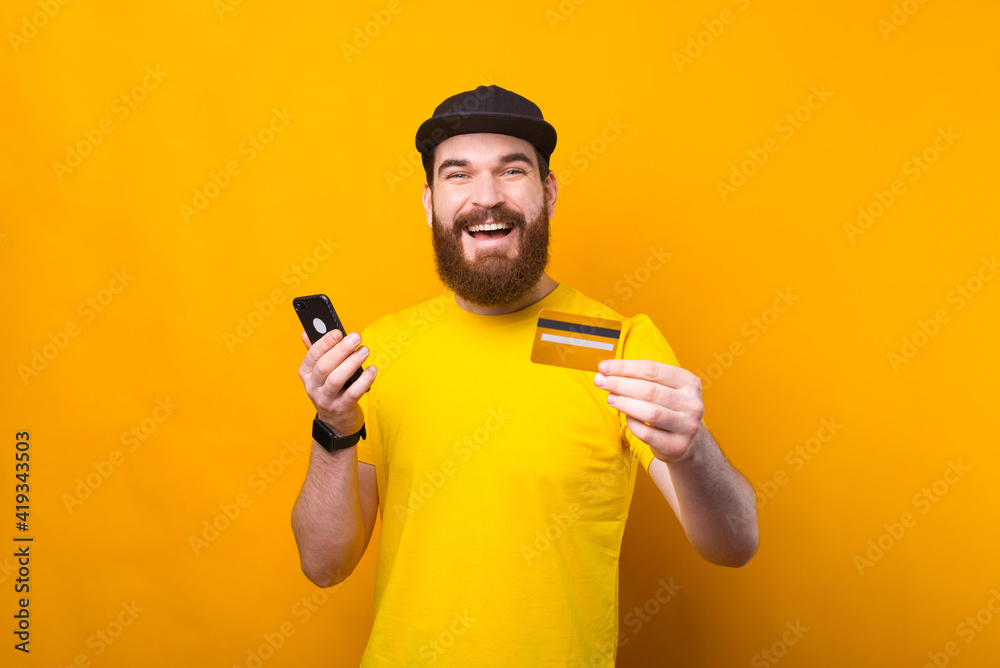 Amazed young bearded man using credit card and smartphone over yellow background Amazed young bearded man using credit card and smartphone over yellow background