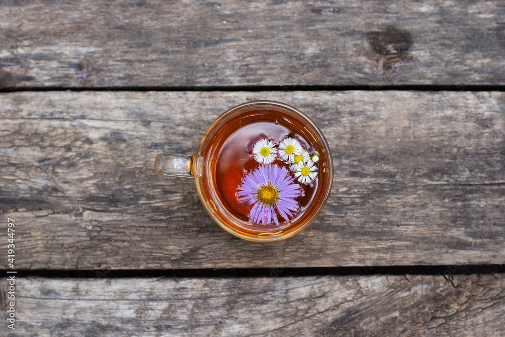 Herbal tea in glass cup with violet and white flowers on wooden background. Top view, copy space. Healthy summer drink. Aster and chamomile. Herbal tea in glass cup with violet and white flowers on wooden background. Top view, copy space. Healthy summer drink. Aster and chamomile.