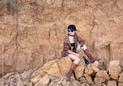 Fashion girl in black crop top, leather boots and brown jacket with bottle of wine and glass on brown dry ground background.