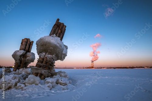 Wooden pillars with blocks of ice stick out from under the ice of a frozen river against the backdrop of a blue sky at sunset.