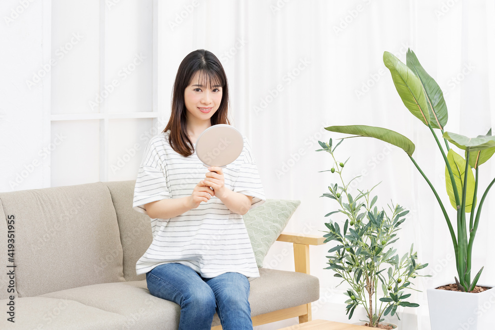 部屋のソファに座って手鏡を使う若い女性