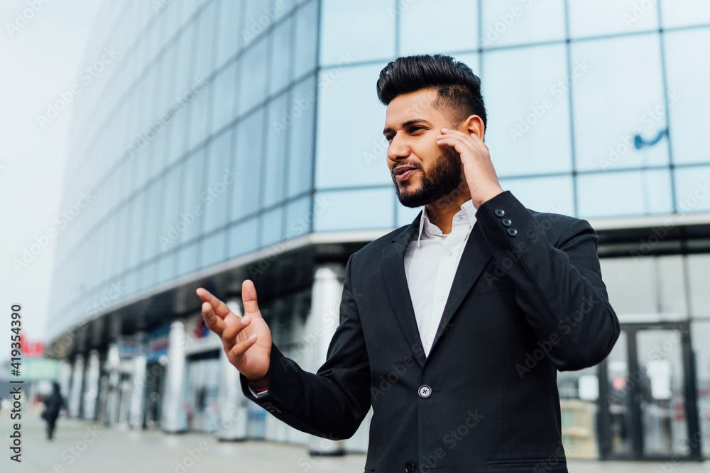 A portrait of an Indian man, an office worker, a bank employee talking on wireless headphones and gesturing, behind him a large modern building A portrait of an Indian man, an office worker, a bank employee talking on wireless headphones and gesturing, behind him a large modern building