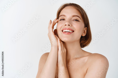 Skin care beauty. Smiling natural woman with naked shoulders and healthy clean and fresh skin, looking happy, touching cheek. Girl apply facial cosmetics, white background