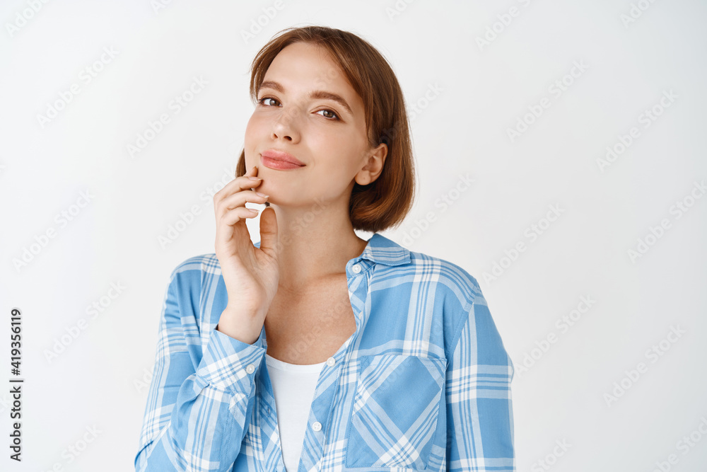 Natural girl touching clean fresh facial skin, glowing nourishing effect on face after daily care product, smiling at camera, standing on white background Natural girl touching clean fresh facial skin, glowing nourishing effect on face after daily care product, smiling at camera, standing on white background