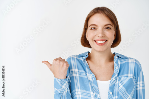 Portrait of pretty female student with white teeth and natural make-up, pointing thumb left and smiling, showing logo brand, standing on white background